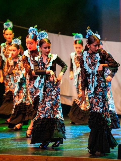Grupo de alumnas de danza infantil con trajes estampados y rosas azules en el pelo, marcando el braceo sobre el escenario